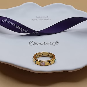 May include: A gold ring with a pink, green, and clear stone design. The ring is on a white plate with the text "Damorecraft Handcrafted Jewelry" on a purple ribbon.