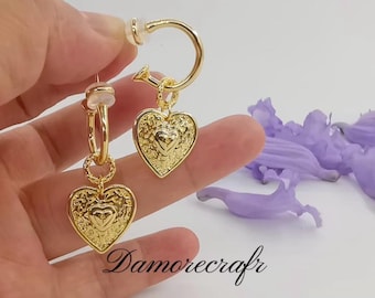 May include: Gold-toned hoop earrings with heart-shaped pendants. The heart pendants have a textured surface with a smaller heart design in the center. The earrings are held by a hand, with purple flower petals in the background. The text "Damorecrafr" is visible.