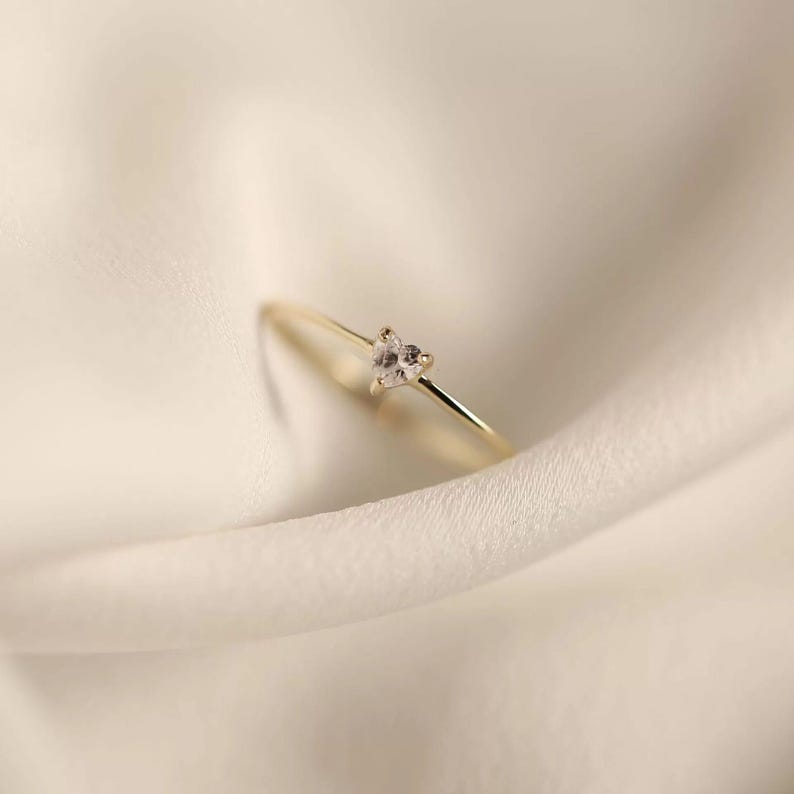 May include: A thin gold band ring featuring a single, small, clear heart-shaped gemstone set in a prong setting. The ring has a polished metallic finish and a delicate, slender profile, showcasing a simple design with a central heart-shaped stone.