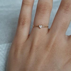 May include: A thin gold band ring featuring a small, clear, heart-shaped gemstone set at the center. The ring has a delicate, minimalist design with a polished metallic finish and a single faceted stone that catches the light.