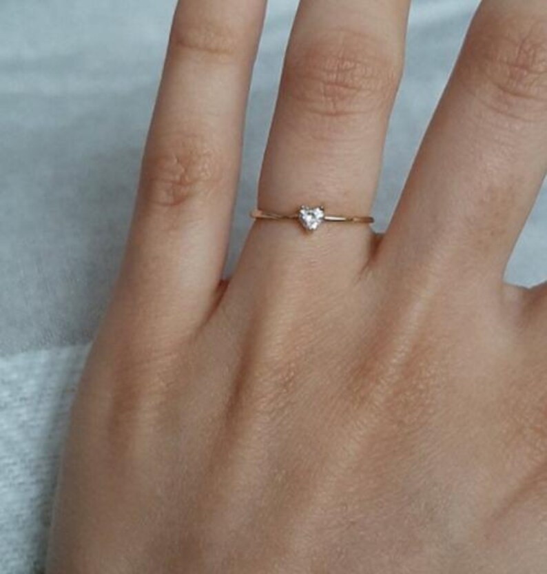 May include: A thin gold band ring featuring a small, clear, heart-shaped gemstone set at the center. The ring has a delicate, minimalist design with a polished metallic finish and a single faceted stone that catches the light.
