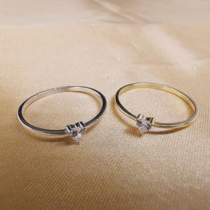 May include: Two thin rings, one silver-toned and one gold-toned, each featuring a small, clear, heart-shaped gemstone set at the center of the band. The rings are simple, delicate, and circular in design, showcasing a polished metallic finish.