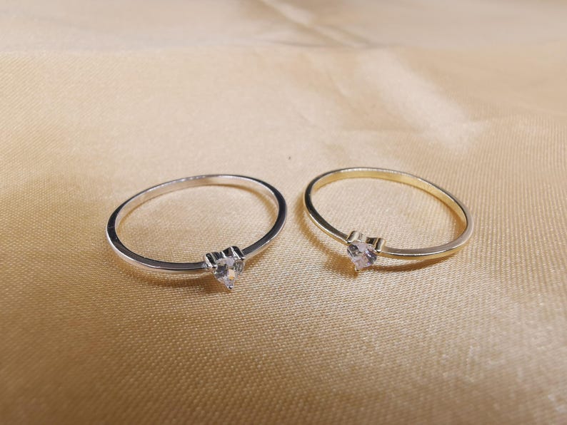 May include: Two thin rings, one silver-toned and one gold-toned, each featuring a small, clear, heart-shaped gemstone set at the center of the band. The rings are simple, delicate, and circular in design, showcasing a polished metallic finish.