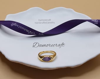 Op de afbeelding: Een gouden ring met een paarse edelsteen op een witte achtergrond. De ring ligt op een klein wit schoteltje met een paars lint waarop "Damorecraft Handmade Jewelry" staat.