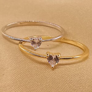 May include: Two thin rings, one in silver-tone metal and one in gold-tone metal, each featuring a small, clear, heart-shaped gemstone set in a prong setting at the center of the band. The rings are positioned overlapping each other.