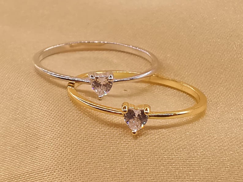 May include: Two thin rings, one in silver-tone metal and one in gold-tone metal, each featuring a small, clear, heart-shaped gemstone set in a prong setting at the center of the band. The rings are positioned overlapping each other.