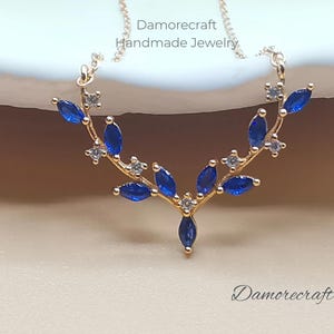 May include: A gold necklace with a delicate branch design featuring blue and white gemstones. The necklace is on a white surface and the text "Damorecraft Handmade Jewelry" is visible above the necklace.