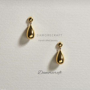 Puede incluir: Un par de pendientes de oro en forma de lágrima con una pequeña bola en la parte superior. Los pendientes están sobre un fondo blanco con el texto "DAMORECRAFT Handcrafted Jewelry" y "Damorecraft" escrito debajo.