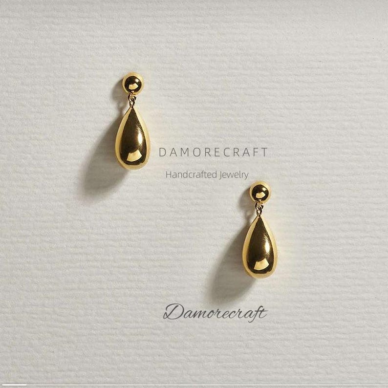 Op de afbeelding: Een paar gouden oorbellen in de vorm van een traan met een kleine bol aan de bovenkant. De oorbellen zijn op een witte achtergrond met de tekst "DAMORECRAFT Handcrafted Jewelry" en "Damorecraft" eronder.
