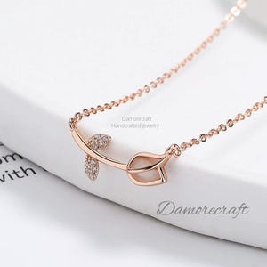 Könnte beinhalten: Eine zarte Halskette aus Roségold mit einem Rosenknospen-Anhänger, der mit einem weißen Stein und funkelnden klaren Steinen besetzt ist. Die Kette ist einfach und besteht aus Gliedern. "Damorecraft Handcrafted Jewelry" ist auf dem weißen Hintergrund gedruckt.