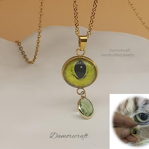 May include: Gold chain necklace with a green eye pendant and a smaller green gemstone pendant. The eye pendant has a black pupil and a yellow iris. The text "Damorecraft Handcrafted Jewelry" is visible in the image.