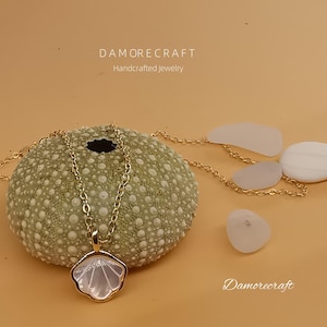 Puede incluir: Un collar de cadena dorada con un colgante de concha blanca. El collar se muestra sobre un erizo de mar y rodeado de vidrio de mar blanco. El texto "DAMORECRAFT Joyería artesanal" es visible en la parte superior de la imagen.