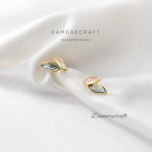 Può includere: Un paio di orecchini a forma di foglia dorati con un design in smalto rosa e blu. Gli orecchini sono realizzati a mano e presentano un delicato design a foglie sovrapposte.