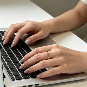 May include: A thin gold ring featuring a single small round clear gemstone set on a slender band. The ring is worn on the ring finger of a hand resting on a silver laptop keyboard with black keys, showcasing a polished metallic finish on the band.