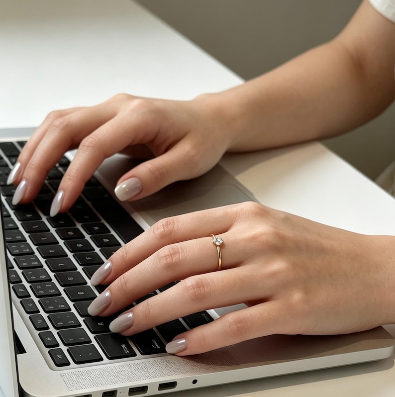 May include: A thin gold ring featuring a single small round clear gemstone set on a slender band. The ring is worn on the ring finger of a hand resting on a silver laptop keyboard with black keys, showcasing a polished metallic finish on the band.