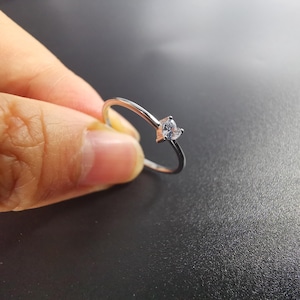 May include: A thin silver-toned metal ring featuring a small, clear, heart-shaped gemstone set at the top. The band is smooth and slender, showcasing a minimalist design with a single faceted stone that catches the light.