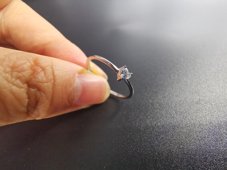 May include: A thin silver-toned metal ring featuring a small, clear, heart-shaped gemstone set at the top. The band is smooth and slender, showcasing a minimalist design with a single faceted stone that catches the light.