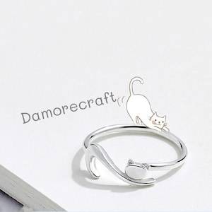 May include: A silver-colored ring with a cat design. The ring features a cat's head and tail forming the band, with a small cat illustration above. The text "Damorecraft" is in the background.