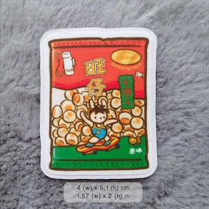 May include: A white sticker with a cartoon illustration of a bag of snacks. The bag is red and green with a cartoon character on a skateboard. The bag has the text "旺仔" in Chinese characters. The sticker is 4 inches wide by 5.1 inches high.