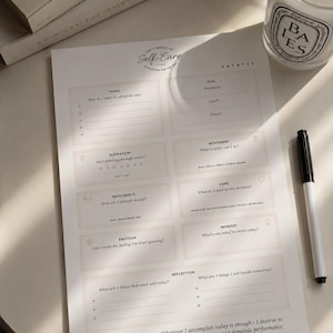 May include: A white self-care planner with sections for tasks, fuel, hydration, movement, indulgence, care, emotion, mindset, and reflection. A black and white pen and a lit candle are next to the planner. A stack of books is in the background.