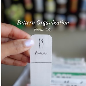 Pode incluir: Uma etiqueta de padrão branca com a etiqueta "Dresses" e uma ilustração simples de um vestido. O texto "Pattern Organization" e "Pattern Tabs" também são visíveis. A etiqueta é segurada por uma mão.