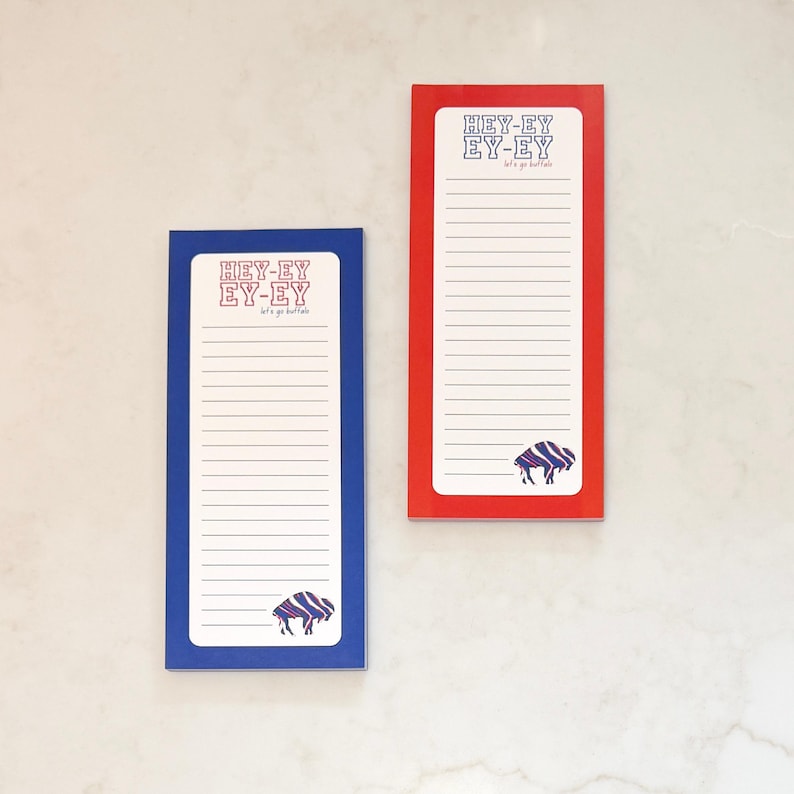 May include: Two rectangular notepads, one blue and one red, with white lined paper. Each notepad features the text "HEY-EY EY-EY" and a buffalo graphic in blue and purple. The notepads are designed for jotting down notes.