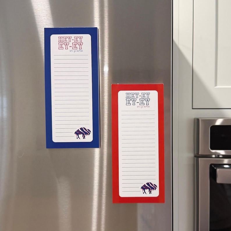 May include: Two rectangular magnetic notepads are displayed on a stainless steel refrigerator. One notepad has a blue border, and the other has a red border. Both have the text "HEY-EY EY-EY let's go buffalo" and a buffalo graphic.