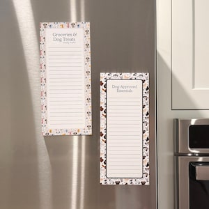 May include: Two rectangular notepads with dog-themed borders are attached to a stainless steel refrigerator. One notepad reads "Groceries & Dog Treats" and the other says "Dog-Approved Essentials". Both have lined pages.
