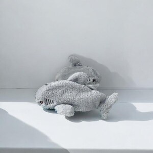 Puede incluir: Dos juguetes de peluche de tibur&oacute;n grises. Los tiburones est&aacute;n tumbados sobre una superficie blanca.