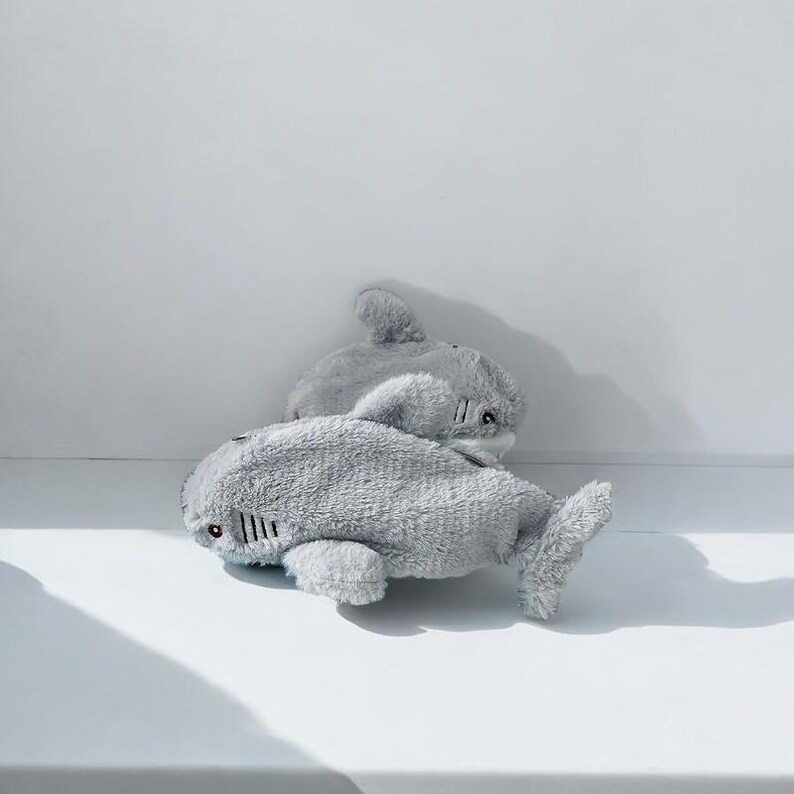 Puede incluir: Dos juguetes de peluche de tibur&oacute;n grises. Los tiburones est&aacute;n tumbados sobre una superficie blanca.
