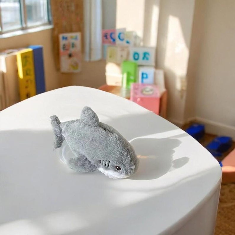 Puede incluir: Un juguete de peluche de tibur&oacute;n gris con vientre blanco y ojos negros. El juguete est&aacute; sobre una superficie blanca.