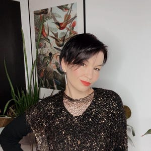 Op de afbeelding: Een vrouw draagt een zwarte top met gouden pailletten en franjes. Ze draagt er een zwart shirt met lange mouwen onder. Een ingelijste prent van kolibries hangt aan de muur achter haar.