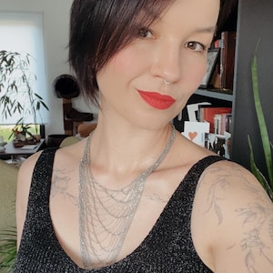 Op de afbeelding: Een mouwloos zwart hemdje met een subtiele metallic glans, gecombineerd met een meerlaagse zilveren ketting. De ketting bestaat uit verschillende delicate, gedrapeerde strengen die vanaf het sleutelbeen naar beneden hangen in een waterval.