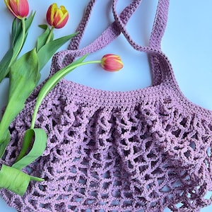 Op de afbeelding: Een lavendelgehaakte draagtas met een netachtig ontwerp en lange banden. De tas wordt getoond met drie tulpen, hun bloemblaadjes een mix van rood en geel, en groene stelen. De achtergrond is licht van kleur.