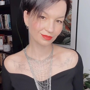 Op de afbeelding: Een vrouw draagt een zwarte off-the-shoulder top en een zilveren ketting. De ketting heeft een geweven, netachtig ontwerp. De vrouw heeft kort, donker haar en rode lippenstift. Ze glimlacht.