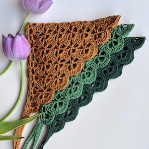 May include: Three crocheted bandanas in shades of brown, green, and dark green, arranged in a layered fan shape. The bandanas have a scalloped edge and a delicate openwork design. Two purple tulips are placed on the left side of the bandanas.