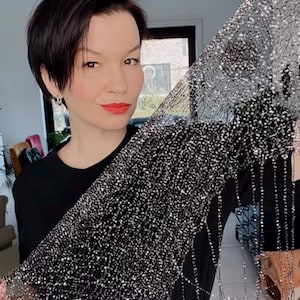 Puede incluir: Se muestra una tela negra, similar a una red, con adornos brillantes. El material está cubierto de pequeños detalles reflectantes, creando un efecto brillante. Este textil decorativo podría usarse para ropa o proyectos de manualidades.