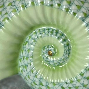 May include: A green and white ceramic seashell with a detailed spiral pattern. The shell has a glossy finish and a gold accent in the center.