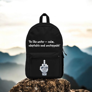 Könnte beinhalten: Schwarzer Rucksack mit einem weißen Zitat, das lautet "Sei wie Wasser - ruhig, anpassungsfähig und unaufhaltsam". Der Rucksack hat eine Tasche auf der Vorderseite mit einer weißen Hand, die den Mittelfinger zeigt, mit einem Om-Symbol auf der Handfläche.