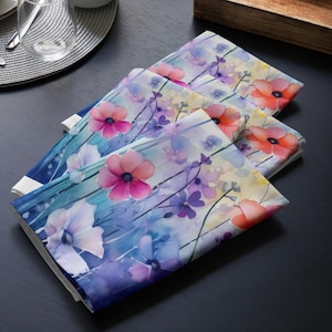 Könnte beinhalten: Set aus vier blauen, violetten und rosafarbenen Blumenservietten mit Aquarellblumen. Die Servietten haben einen weißen Rand und zeigen verschiedene Blumen, darunter Anemonen und Mohnblumen.