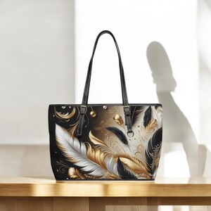 Könnte beinhalten: Schwarze Tragetasche mit einem Federdesign in Gold, Weiß und Schwarz. Die Tasche hat schwarze Griffe und einen schwarzen Rand. Die Tasche steht auf einer Holzoberfläche.
