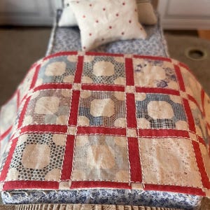 May include: A miniature bed featuring a patchwork quilt with a grid of red fabric strips separating squares of cream, blue, and white patterned fabric. A small cream pillow with red polka dots rests at the head of the bed, which is covered in blue fabric.