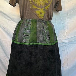 May include: A brown t-shirt with a yellow sun and skull graphic, paired with a knee-length skirt. The skirt features a green and black patchwork design with a green trim at the hem. The skirt is approximately 28 inches long.