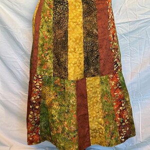 May include: A patchwork skirt with vertical panels in shades of green, yellow, brown, and rust. The skirt features floral and leaf patterns, creating a bohemian aesthetic. The skirt is displayed on a black mannequin base.
