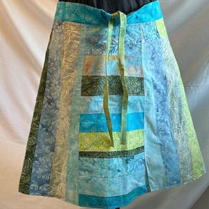May include: A patchwork skirt in shades of blue, green, and orange. The skirt features a drawstring waist and is made of various fabric panels with floral and abstract patterns. The skirt is displayed on a mannequin.