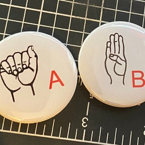May include: Three white round buttons with black and white illustrations of hands signing the letters A, B, and C. The letters are in red.