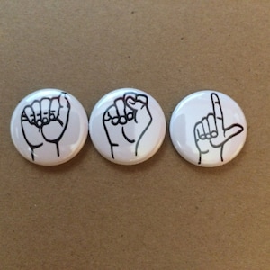 May include: Three white round buttons with black outlines of hands in different sign language positions.