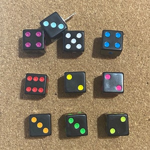 May include: Nine black dice with colored dots, including pink, white, blue, red, yellow, and orange. One die has a push pin through it. The dice are arranged on a brown corkboard surface.