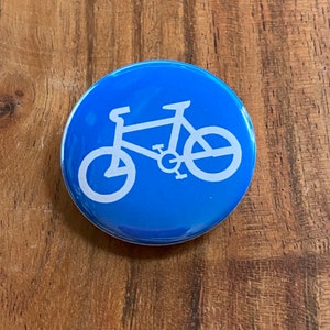 Könnte beinhalten: Ein blauer Knopf mit einer weißen Silhouette eines Fahrrads. Das Fahrrad hat ein einfaches Design mit zwei Rädern, einem Lenker und einem Sattel.