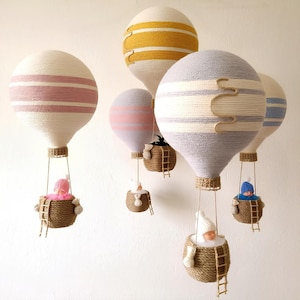 May include: Five handmade hot air balloons with striped patterns in pink, yellow, and gray. Each balloon has a small basket with a doll inside. The balloons are suspended from the ceiling.